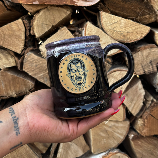 Handmade 14oz black stoneware mug with 2024 Darkest Day Folly Vampire emblem held in hand against stacked firewood background